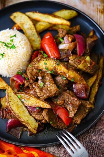 An overhead view of a serving of Classic Lomo Saltado, Peruvian Steak and Potatoes Stir-Fry, on a black dinner plate with a mound of Peruvian Garlic Rice on the plate as well.