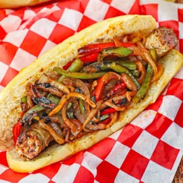 An overhead view of a NYC-Style Sausage and Peppers nestled into a homemade Italian roll resting in a basket lined with a red-checkered piece of wax paper.