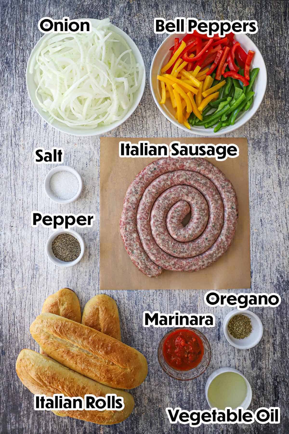 An arrangement of ingredients for Sausage and Peppers on a grey wooden background including a rope of uncooked Italian sausage, onion, bell peppers, Italian rolls, marinara sauce, oil, and seasonings.