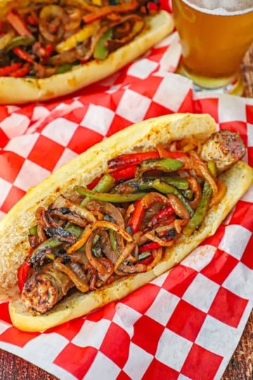 A close-up view of a NYC-Style Sausage and Peppers in a homemade Italian roll resting in a basket lined with red-checkered wax paper.