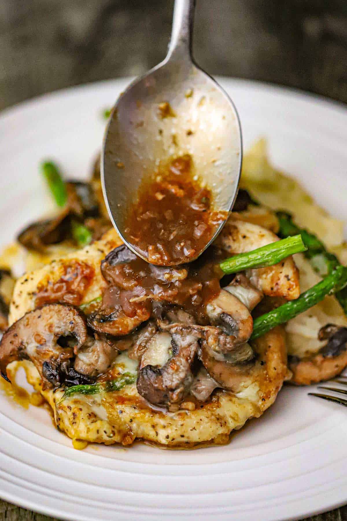 A spoon being used to drizzle a brown sauce over the top of a serving of chicken Madeira with mushrooms and asparagus on a white dinner plate.