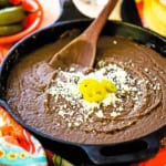 A close-up view of a large black cast-iron skillet filled with authentic homemade refried beans topped with crumbled Cotija cheese and three slices of pickled jalapeños.