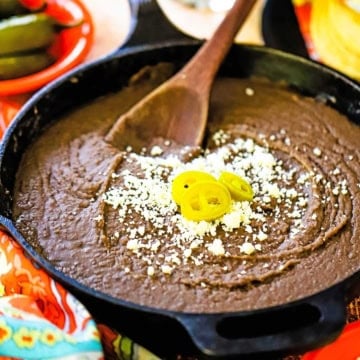 A close-up view of a large black cast-iron skillet filled with authentic homemade refried beans topped with crumbled Cotija cheese and three slices of pickled jalapeños.