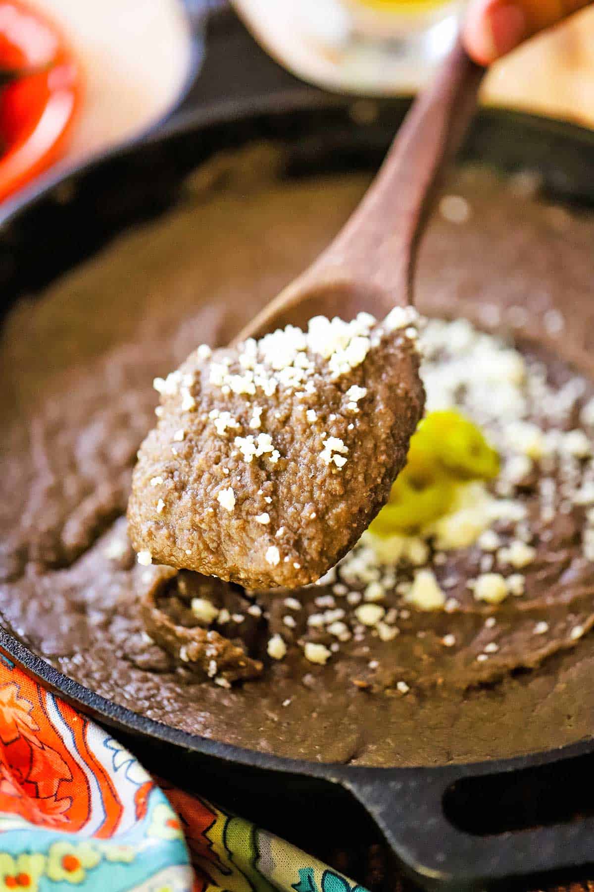 A person using a large wooden spoon to lift up a serving of homemade authentic refried beans from a skillet filled with the same.