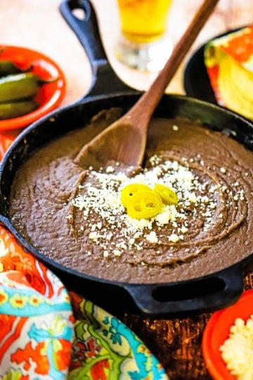 A large black cast-iron skillet filled with authentic homemade refried beans topped with crumbled Cotija cheese and three slices of pickled jalapeños.