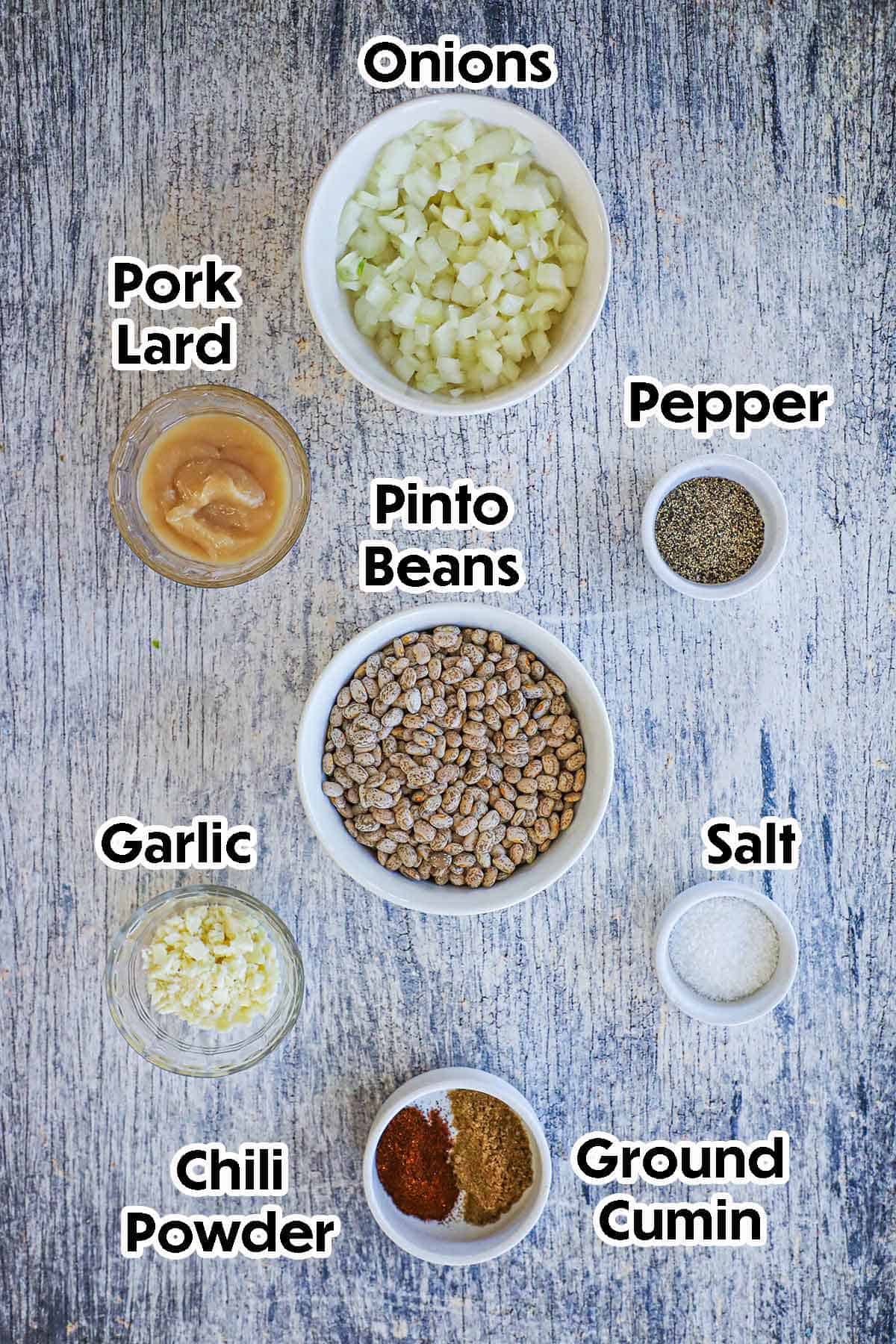 An arrangement of ingredients for homemade authentic refried beans on a grey wooden background including dried pinto beans, chopped onions, pork lard, garlic, and seasonings.