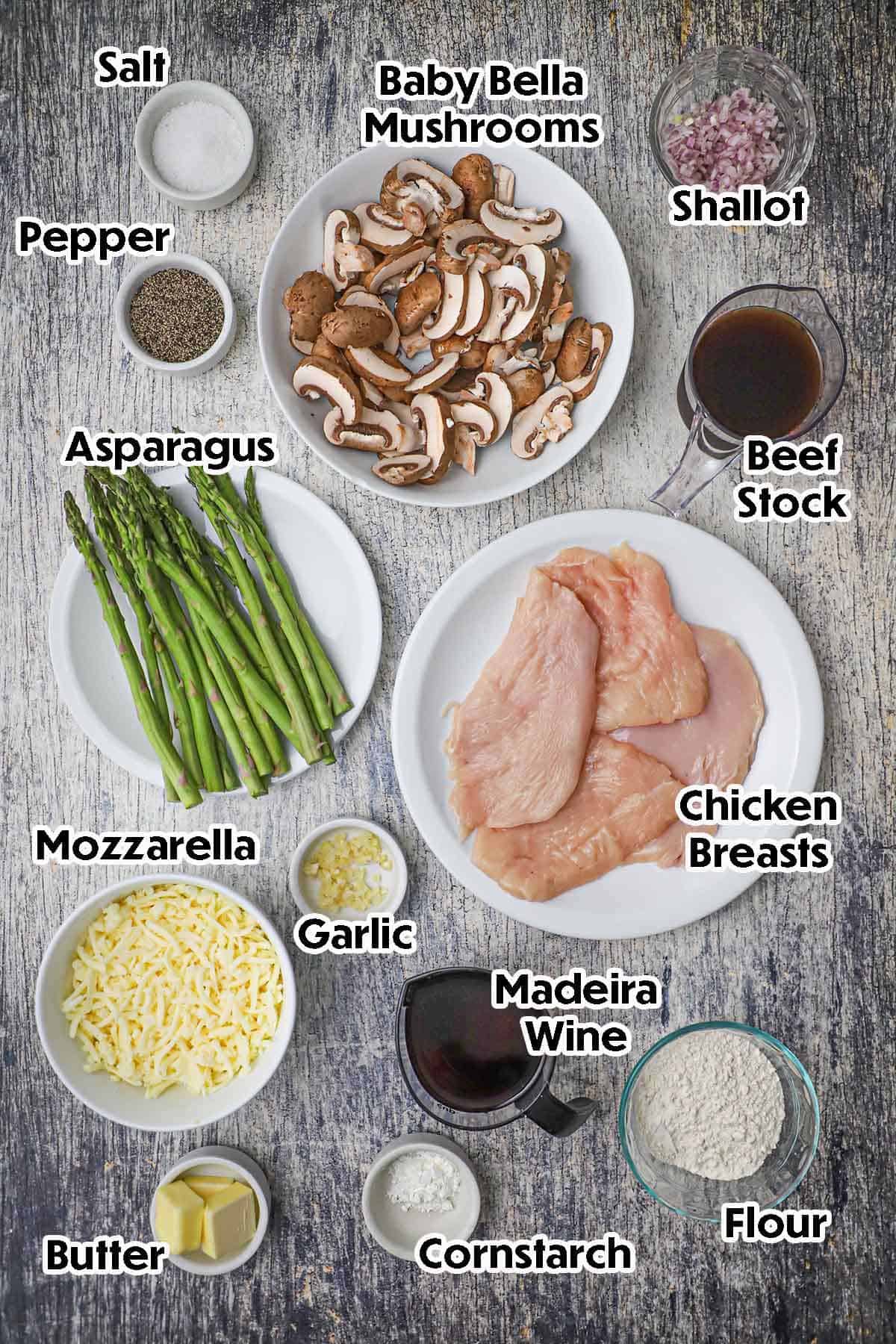 An arrangement of ingredients for Chicken Madeira on a grey wooden background including chicken breast, mushrooms, asparagus, garlic, shallot, beef stock, Madeira wine, flour, Mozzarella cheese, and seasonings.
