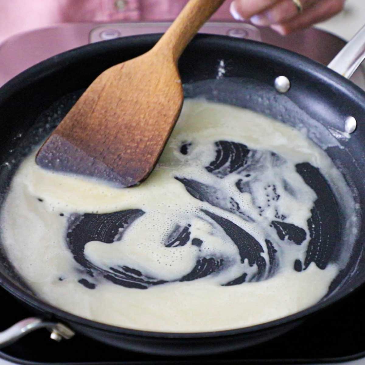 A person using a wooden spatula to stir flour in hot oil in a skillet to create a roux.