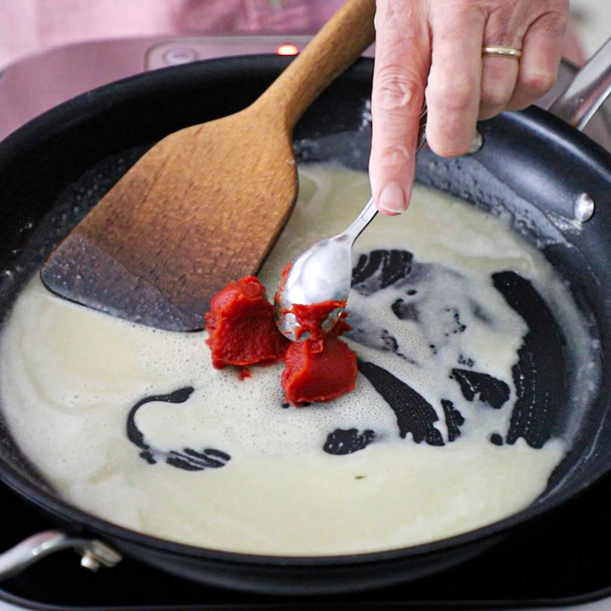 A person adding two tablespoons of tomato paste into a skillet that has a simmering roux in it.