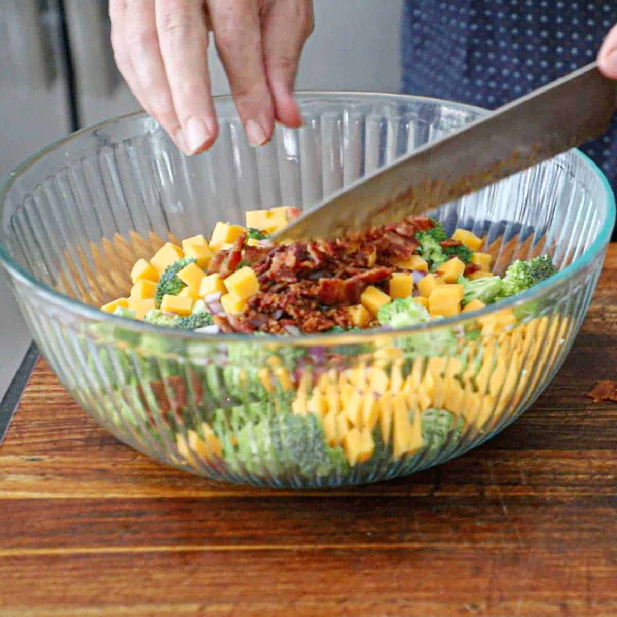 A person using a large chef's knife to transfer cooked crumbled bacon from a cutting board into a large glass bowl filled with broccoli florets, cubed cheddr cheese, and diced red onion.