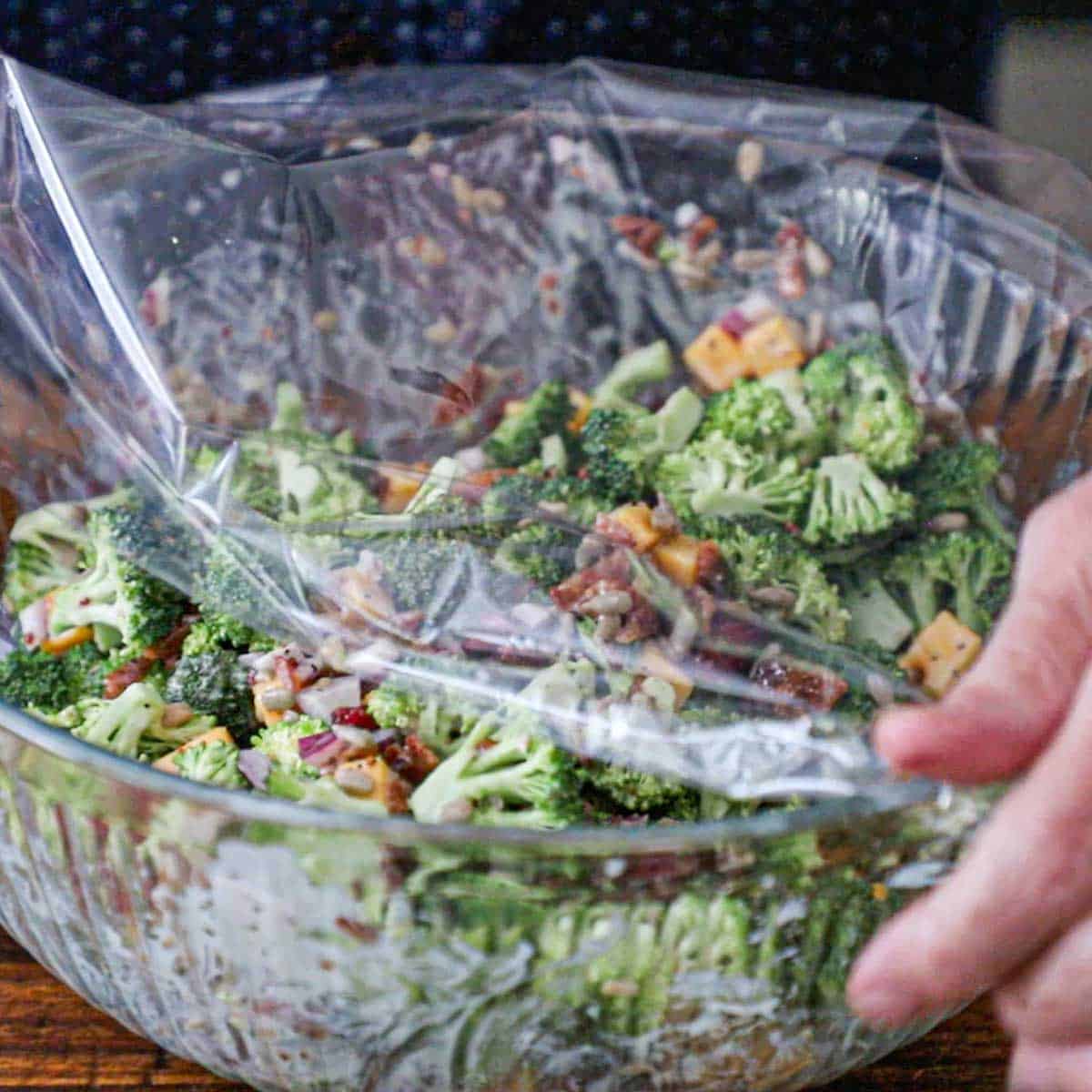 A person plastic plastic wrap over the top of a large glass bowl filled with a freshly prepared broccoli salad with bacon.