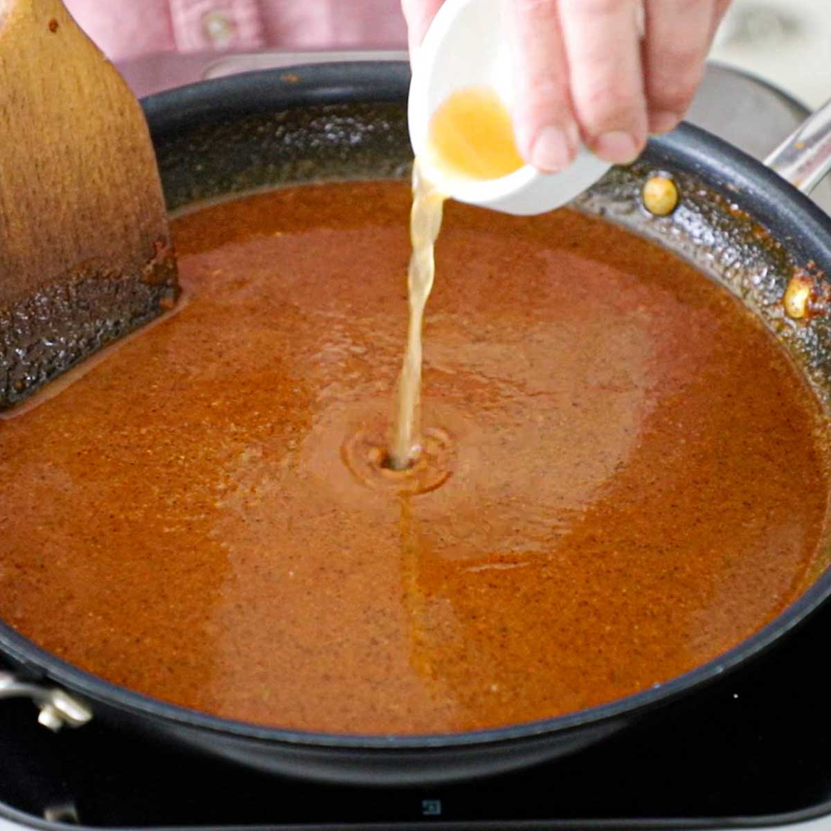 A person pouring apple cider vinegar from a small white bowl into a skillet filled with simmering Easy Homemade Red Enchilada Sauce