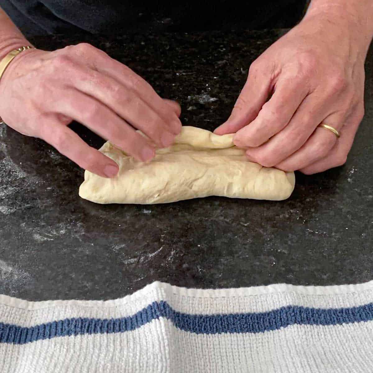 A person folding over the edges of a rectangular piece of Italian rolls dough on a black marble countertop.