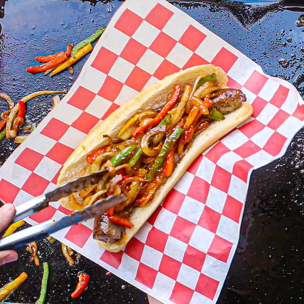 A person using a pair of metal tongs to place sautéed onions and peppers over the top of a cooked link of Italian sausage inside an opened Italian roll.