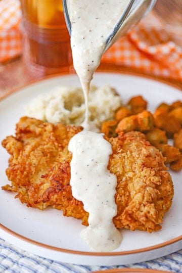 A person pouring cream gravy over a cutlet of Southern chicken fried chicken on a white dinner plate with fried okra.
