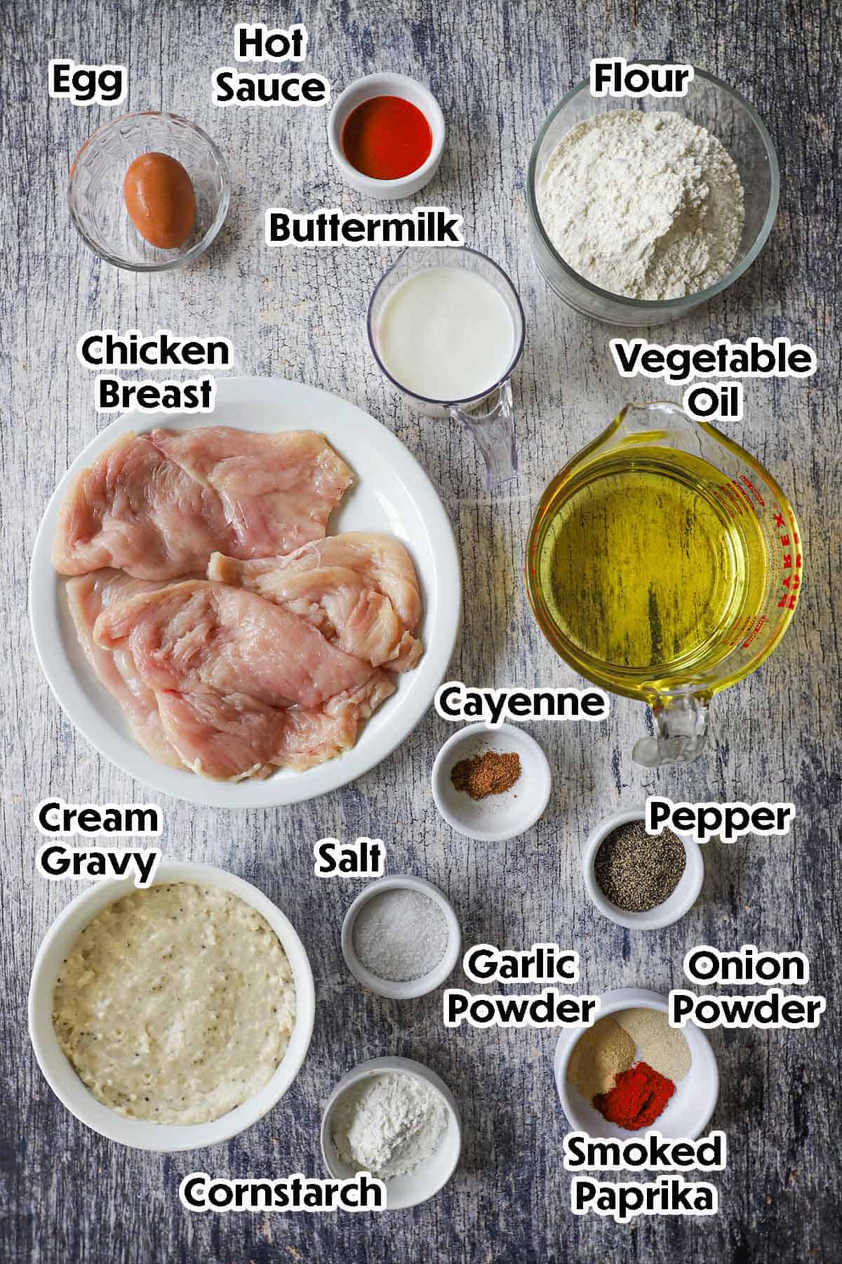 An arrangement of ingredients for Southern Chicken Fried Chicken on a grey wooden background including thin chicken breast, flour, an egg, buttermilk, oil, seasonings, and cream gravy.