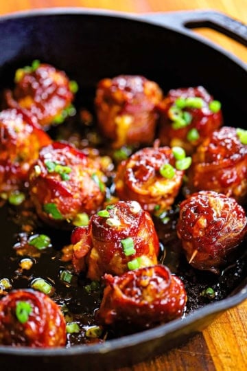 A cast-iron skillet filled with smoked bacon-wrapped meatballs with cheddar and jalapeño, topped with chopped scallions.