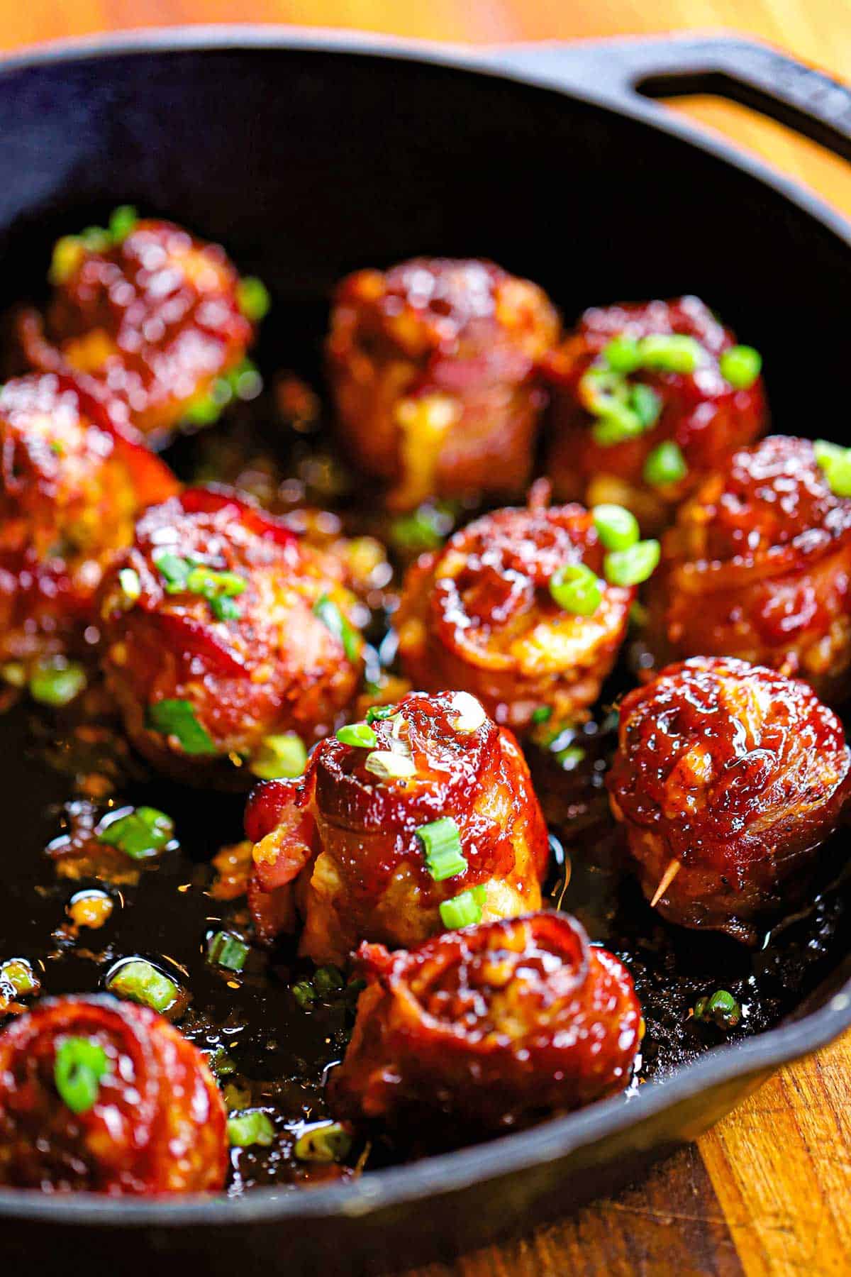 A cast-iron skillet filled with smoked bacon-wrapped meatballs with cheddar and jalapeño, topped with chopped scallions.
