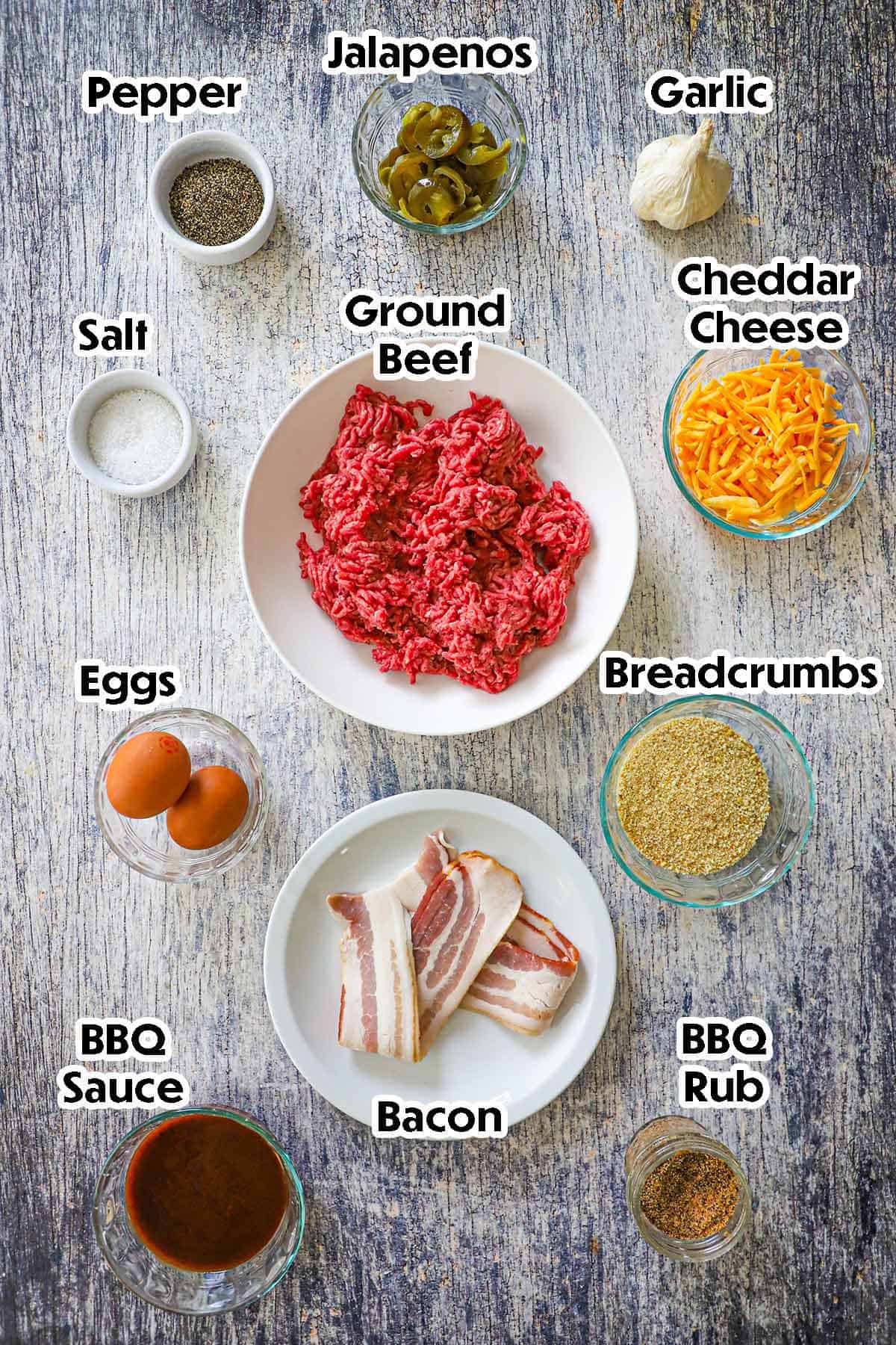 An arrangement of ingredients for Smoked Bacon-Wrapped Meatballs on a grey wooden background including ground beef, breadcrumbs, cheese, eggs, garlic, jalapeños, bacon, BBQ sauce, and seasonings.