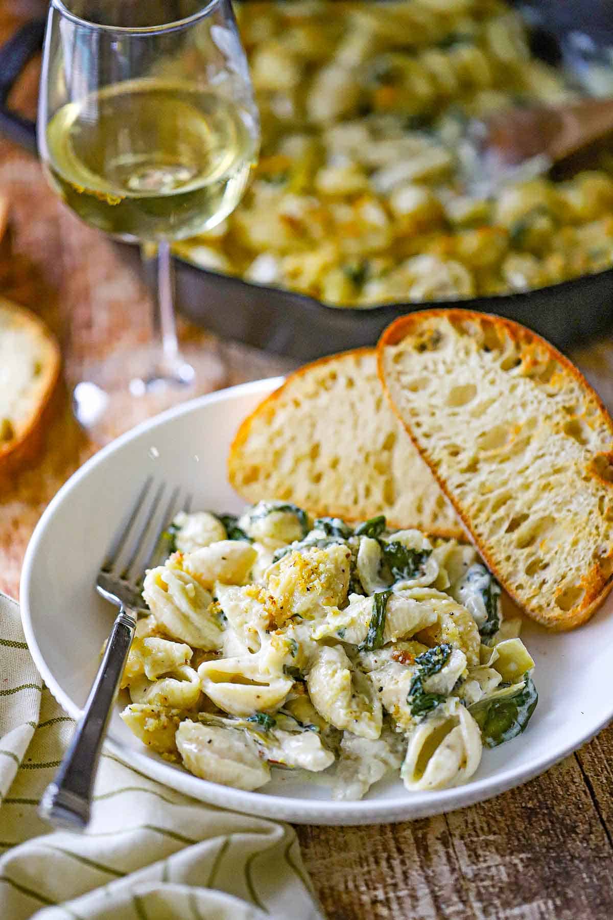 A white pasta bowl filled with a serving of Spinach Artichoke Dip Pasta with two slices of toasted Italian bread resting next to the pasta.