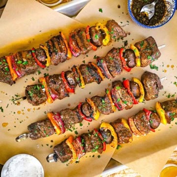 An overhead view of four metal skewer filled with grilled steak kabobs of chunks of beef tenderloin, vegetables, and mushrooms topped with a garlic and balsamic vinaigrette.