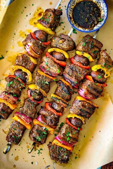 An overhead view of four metal skewer filled with grilled steak kabobs of chunks of beef tenderloin, vegetables, and mushrooms topped with a garlic and balsamic vinaigrette.