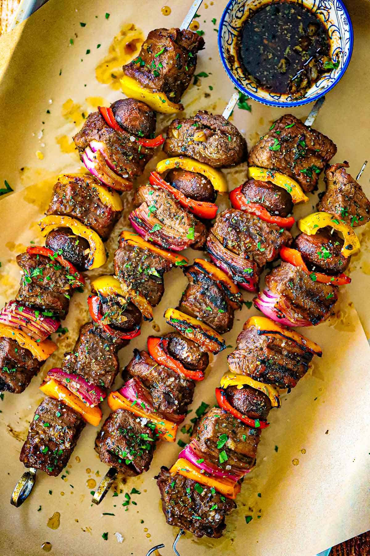 An overhead view of four metal skewer filled with grilled steak kabobs of chunks of beef tenderloin, vegetables, and mushrooms topped with a garlic and balsamic vinaigrette.