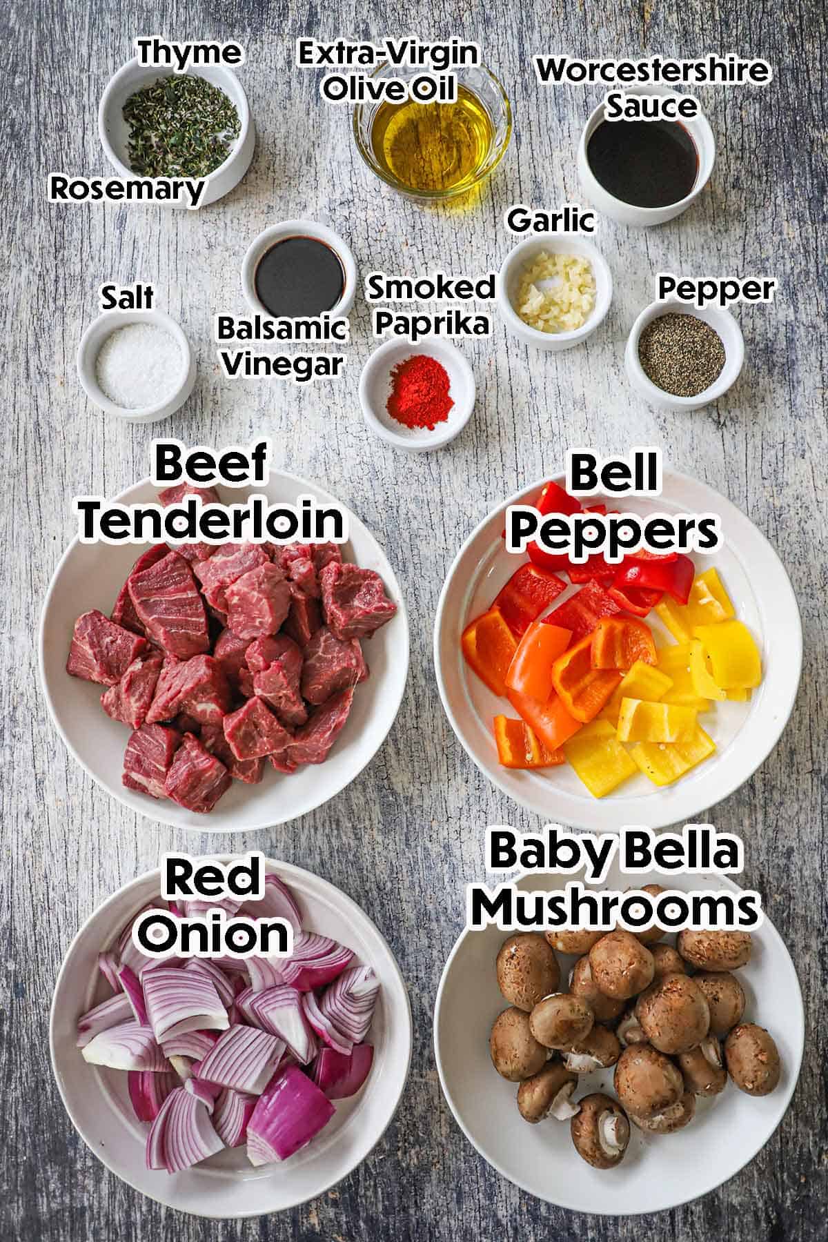 An arrangement of ingredients for grilled steak kabobs on a grey wooden background including beef tenderloin, bell peppers, onion, baby Bella mushrooms, Worcestershire sauce, balsamic vinegar, garlic, herbs, and spices.