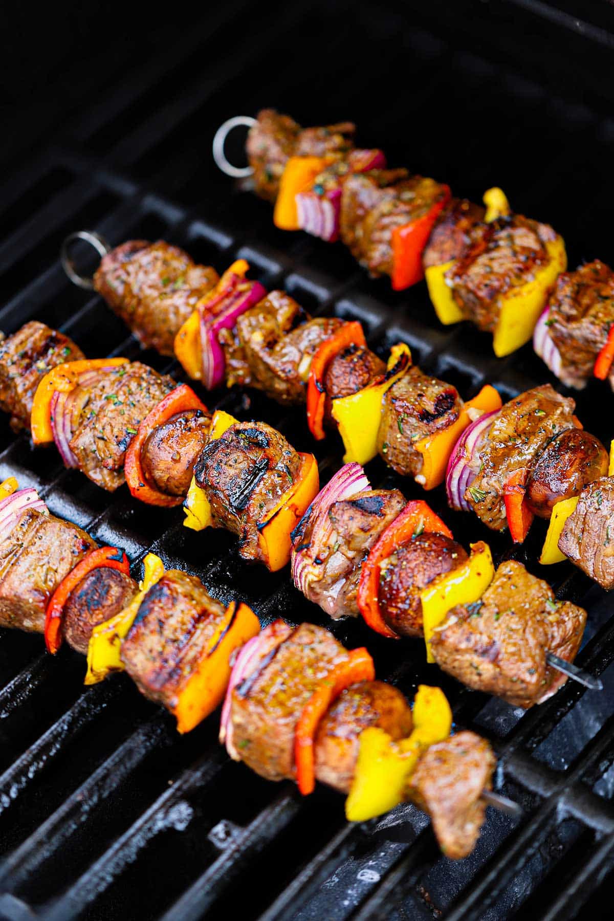 Four metal skewers filled with lightly charred steak kabobs that are being grilled on a gas stove.