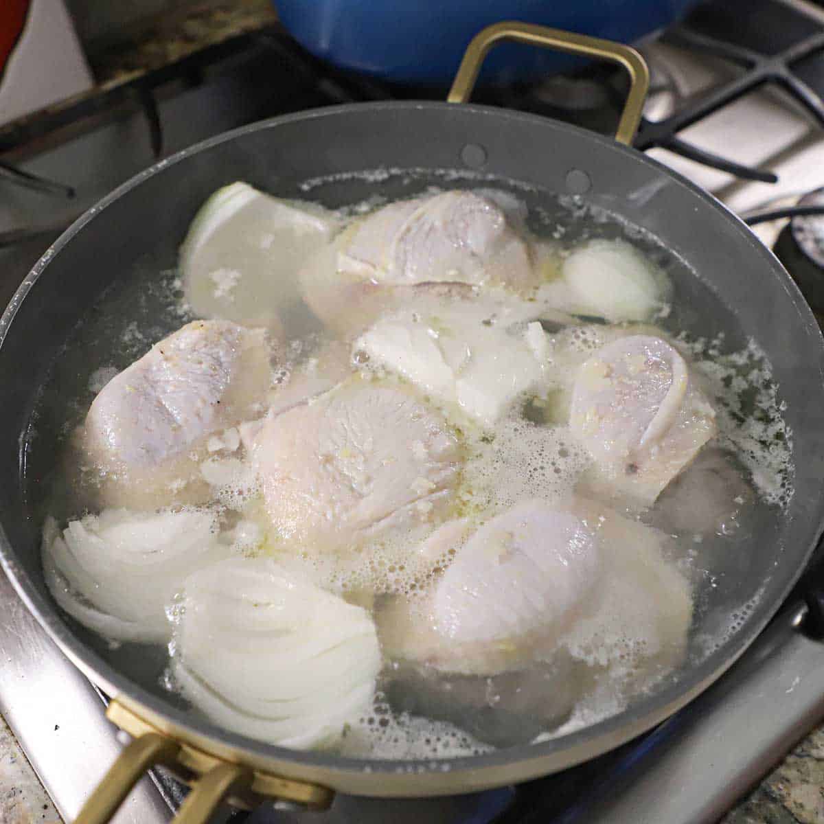 A large skillet filled with skin-on chicken thighs and wedges of onion being poached in water on a gas stove.