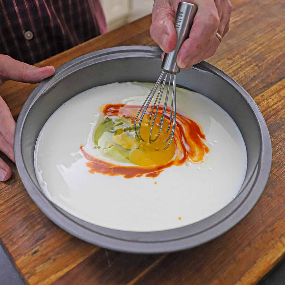 A person using a small whisk to mix an egg and hot sauce into buttermilk in a round baking pan.