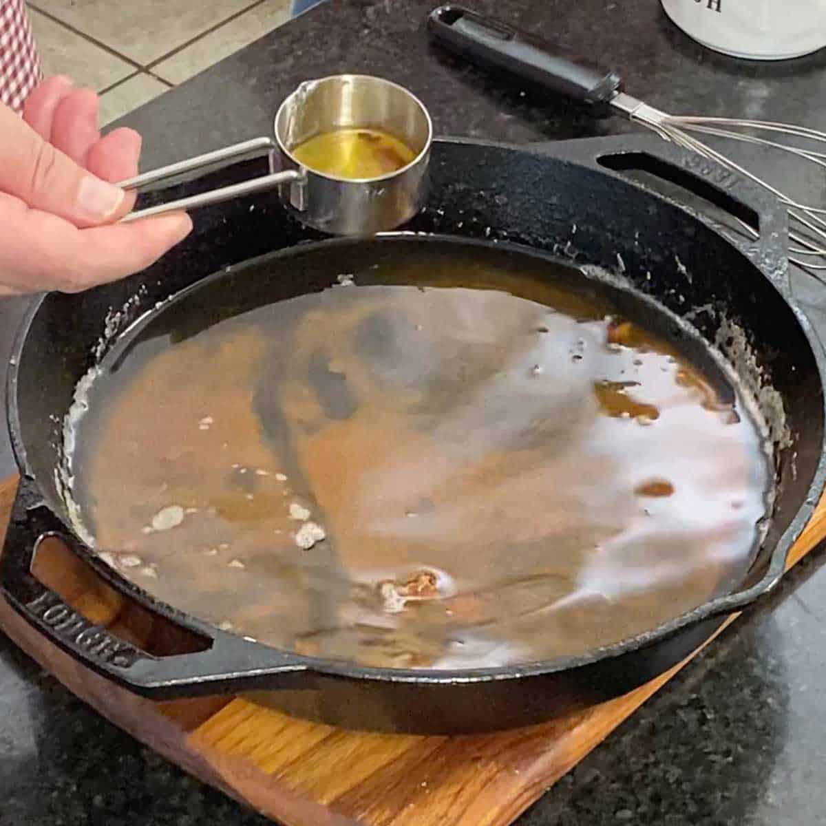 A person holding a metal quarter-cup measuring cup that is partially filled with pan drippings from a skillet below that is filled with oil that was used to fry chicken.