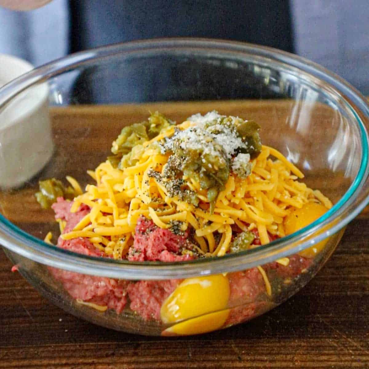 A glass bowl filled with ground beef, two eggs, shredded cheddar cheese, chopped pickled jalapeños, and seasonings.
