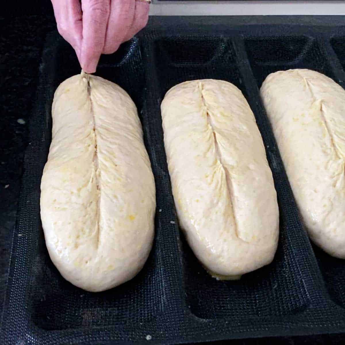 A person using a razor blade to score loaves of risen, un-baked homemade Italian rolls dough that are resting in the channels of an aerated silicone baking mat.