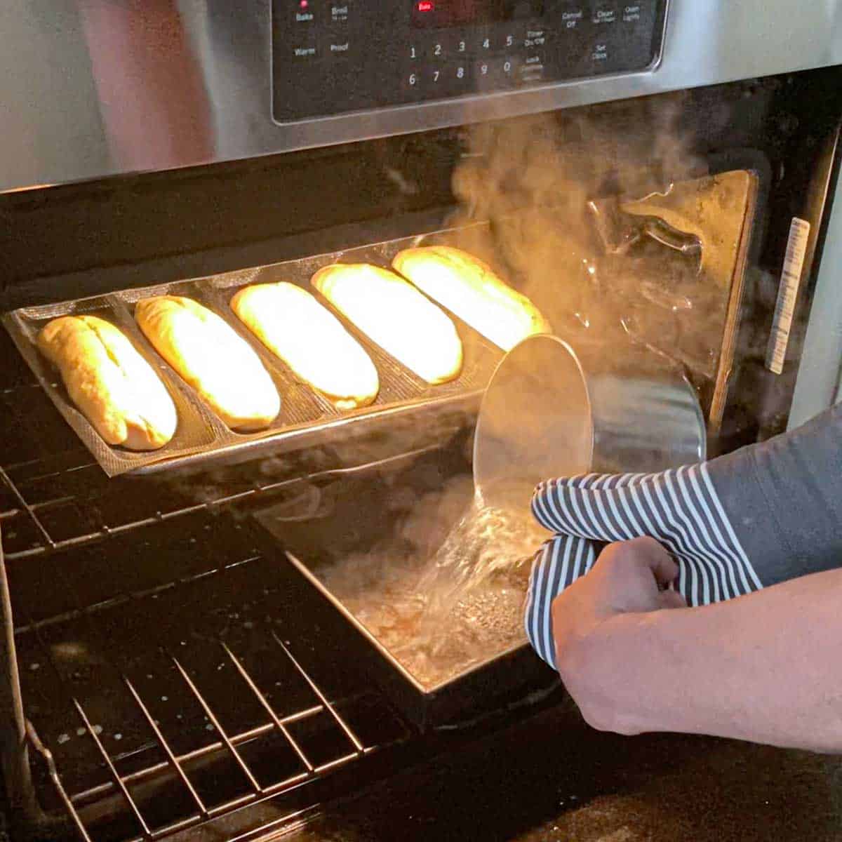 A person transferring boiling water from a pan into a baking pan that is an oven with a row of un-baked homemade Italian rolls on the grate above it.