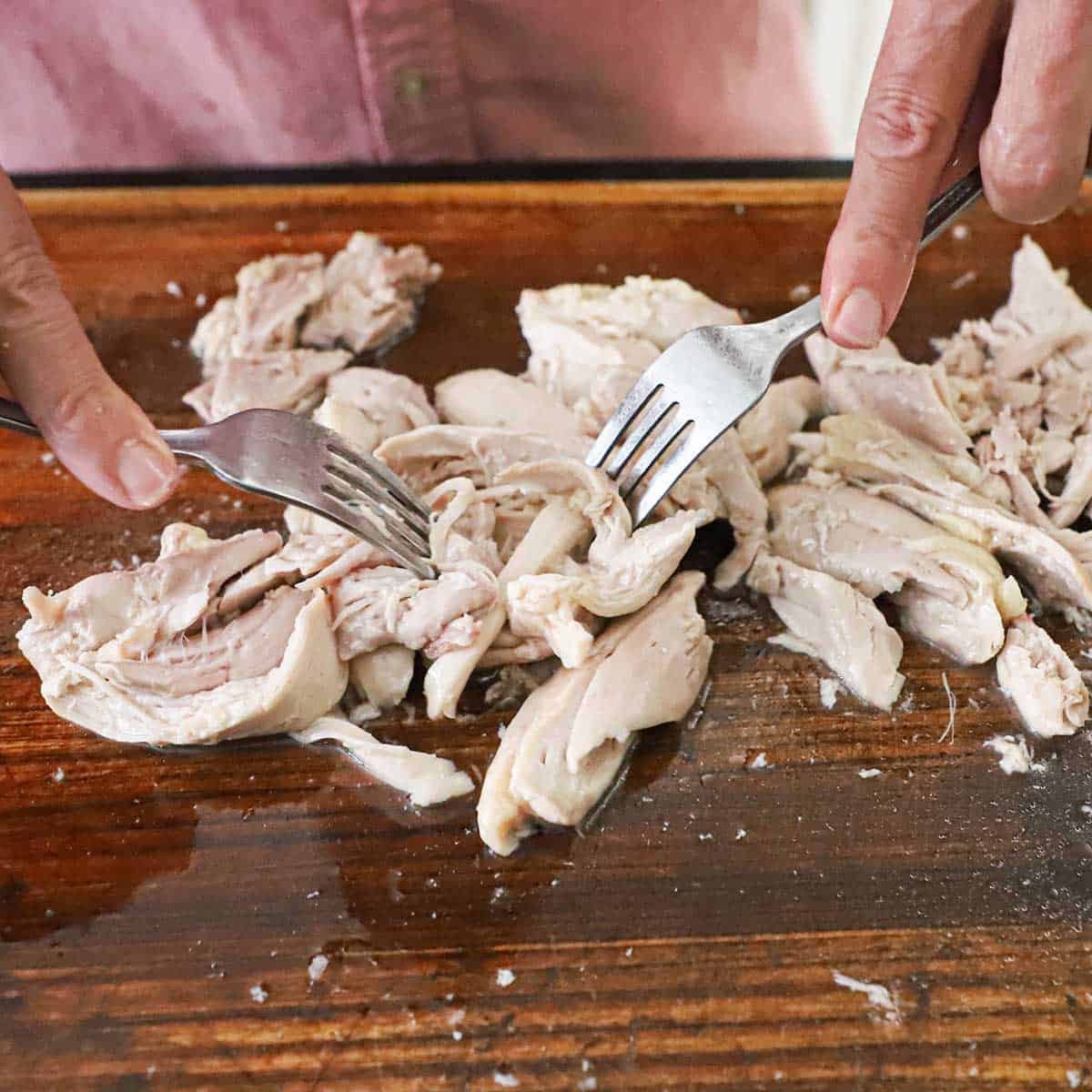 A person using two forks to shred poached chicken thighs on a wooden cutting board.