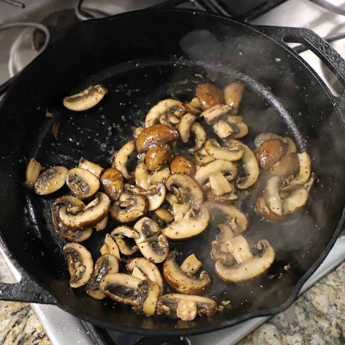 Sliced baby bella mushrooms being sautéed in butter in a large black cast-iron skillet on a gas stove.