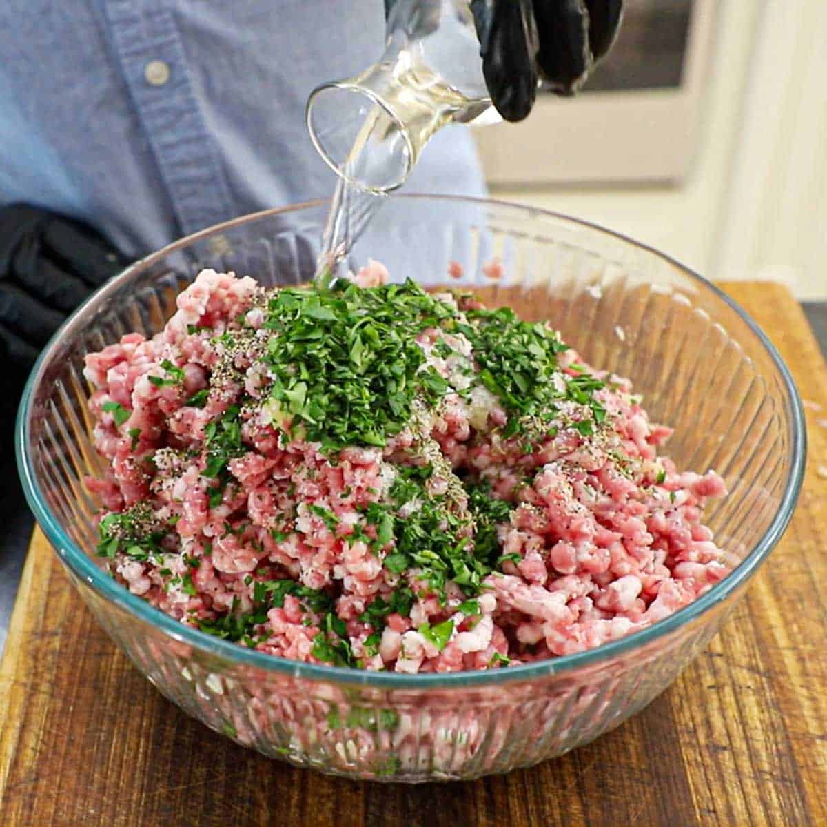 A person pouring white wine from a small carafe into a large glass bowl filled with ground pork butt, Italian parsley, and other Italian sausage seasonings.