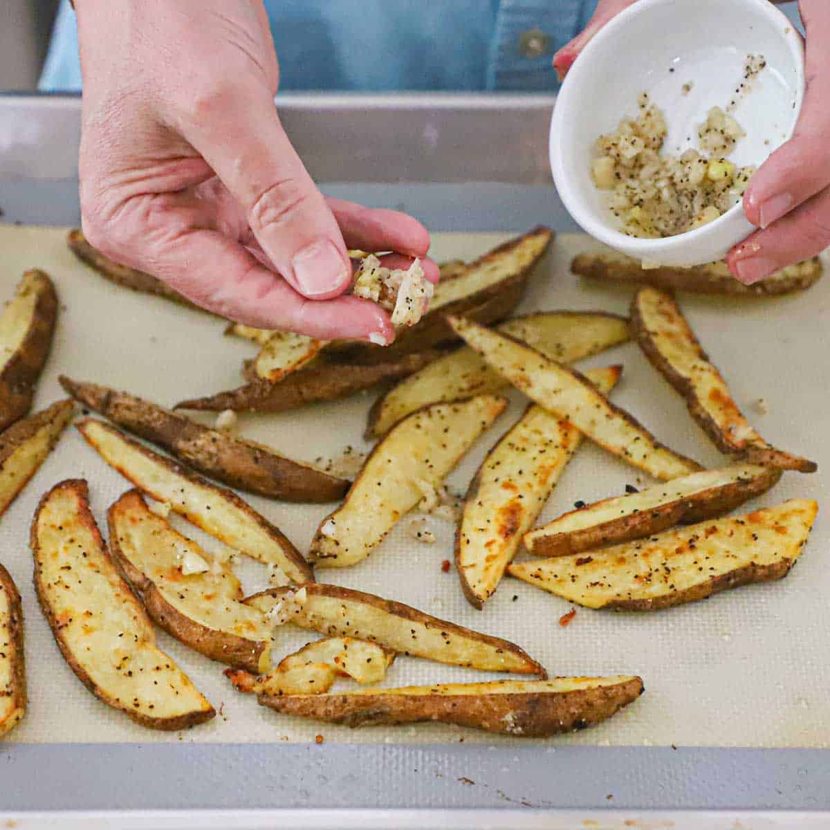 A person sprinkling minced garlic over bake steak fries that are on a silicone mat on a baking pan.
