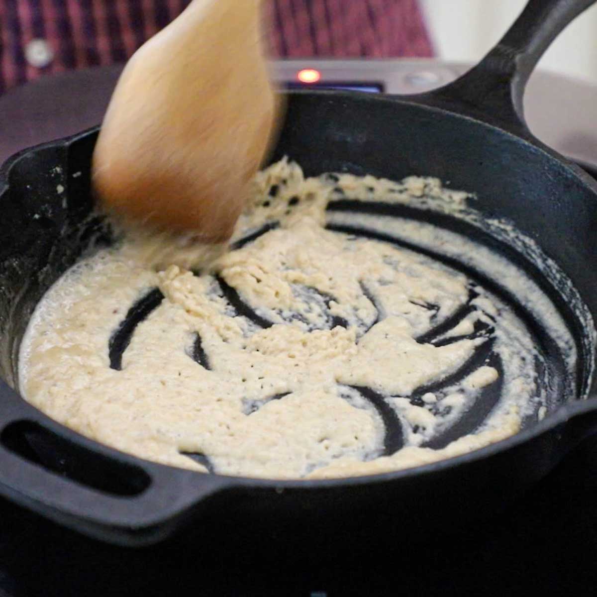 A person using a wooden spoon to stir a roux of flour and fried chicken pan drippings in a black cast-iron skillet.