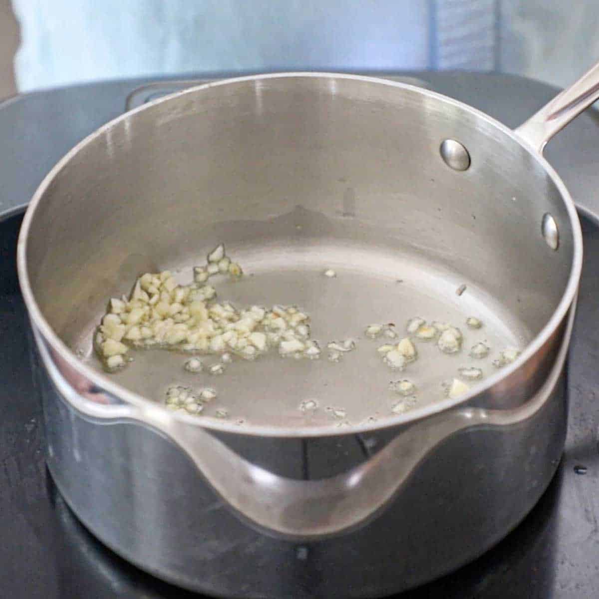 A silver saucepan on a stovetop containing minced garlic that is being sautéed in olive oil.