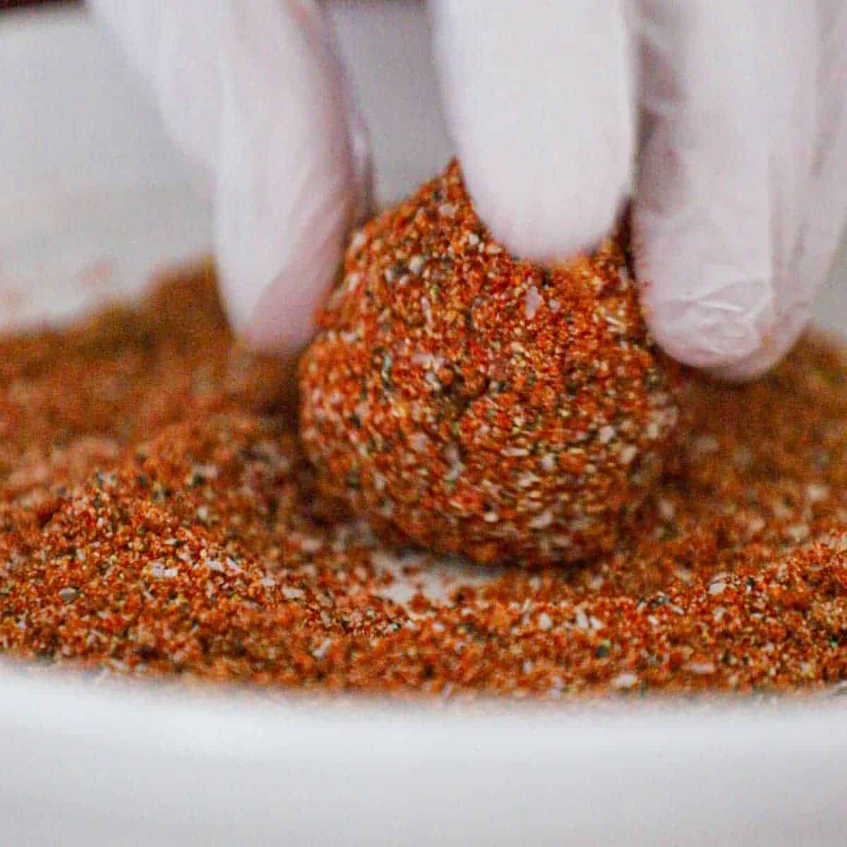 A person rolling an uncooked meatball in a BBQ rub that is in a shallow bowl.