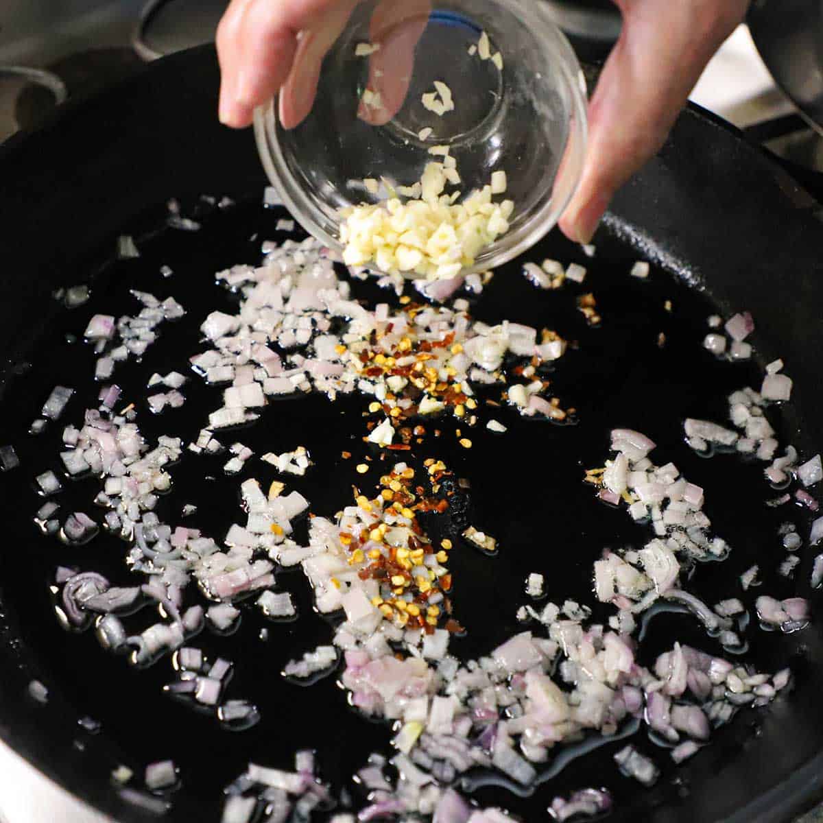 A person transferring minced garlic from a small glass bowl into a cast-iron skillet that is filled with sautéed shallots and red pepper flakes.