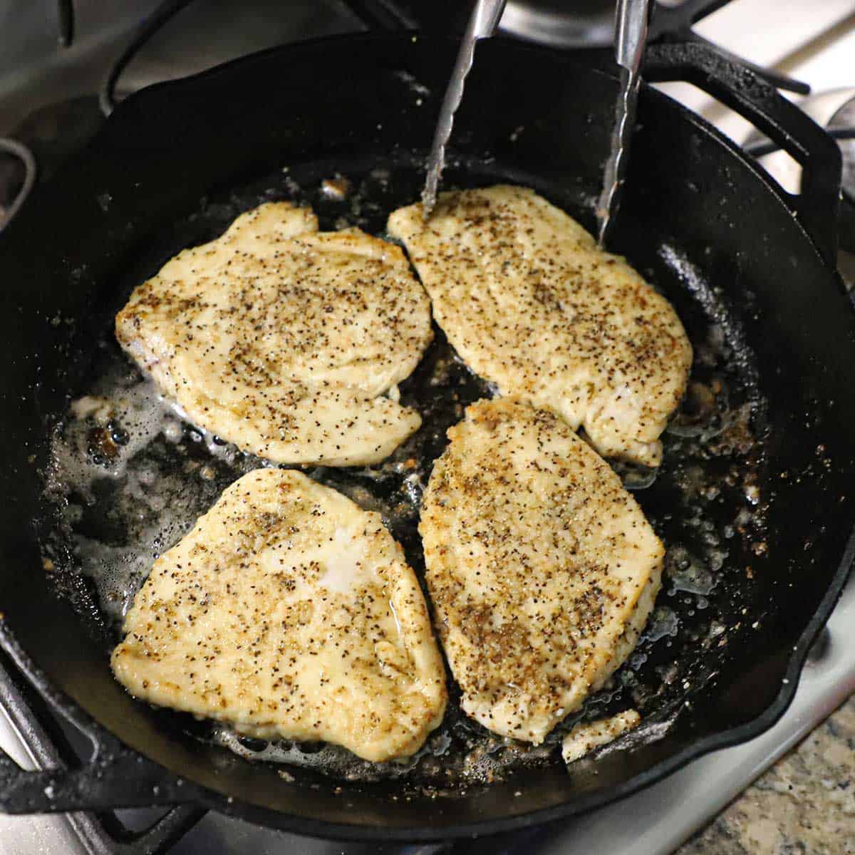 Four seasoned thin chicken breasts being seared in butter in a large black cast-iron skillet on a gas stove.