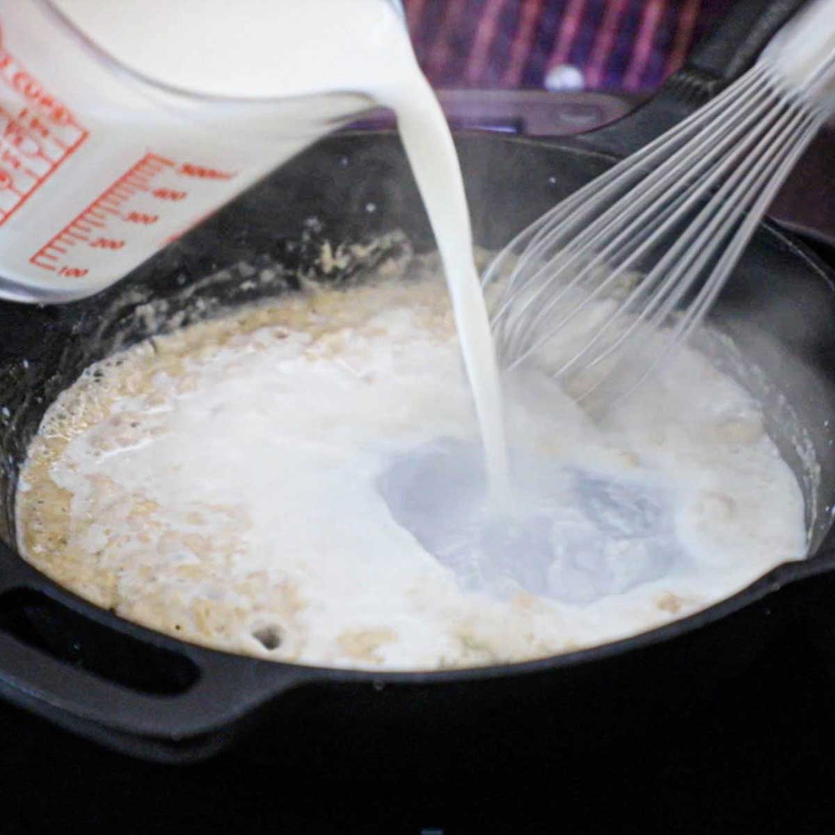 A person whisking whole milk into a roux in a cast-iron skillet.