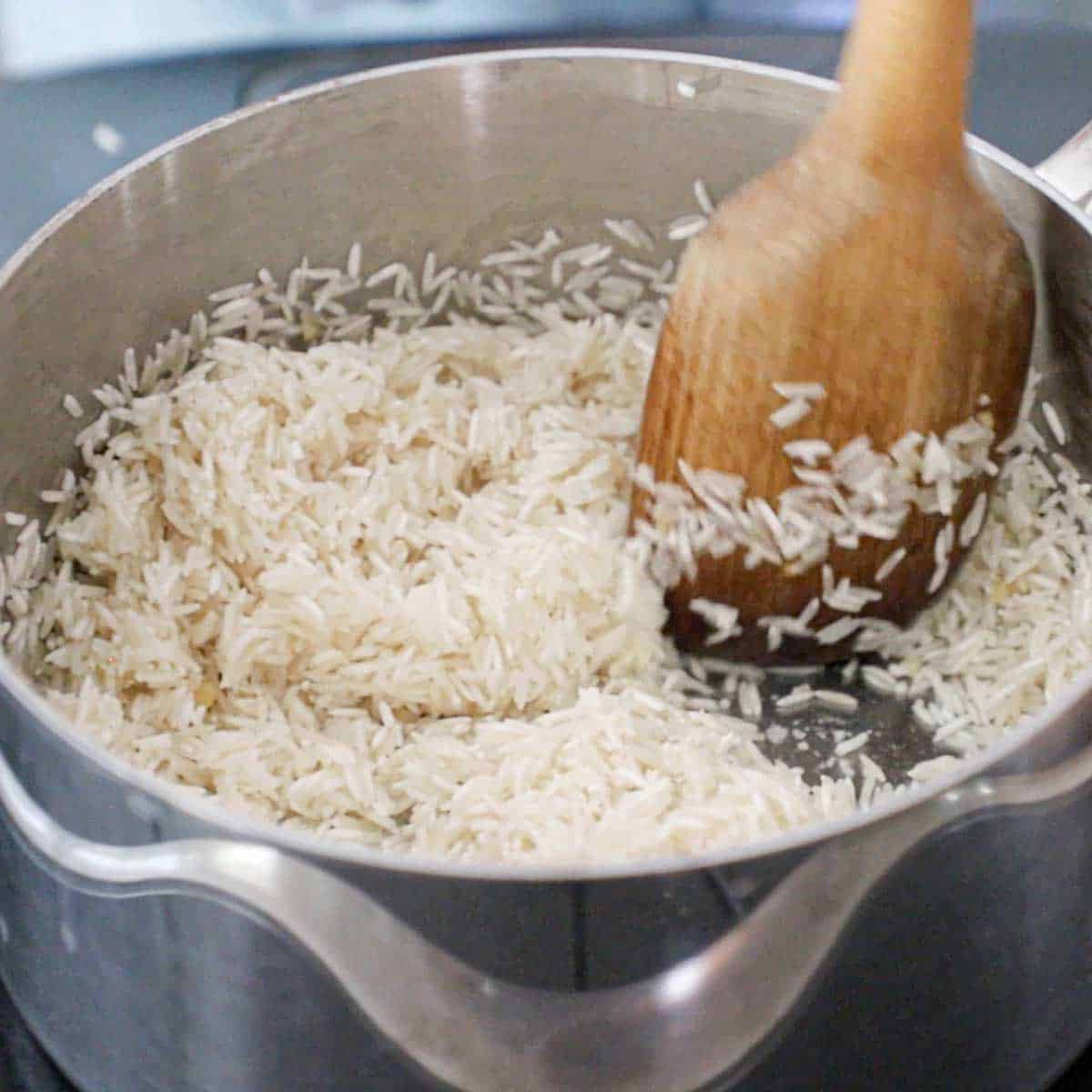 A person using a large wooden spoon to cook Jasmine rice in oil in a silver saucepan on a stove.