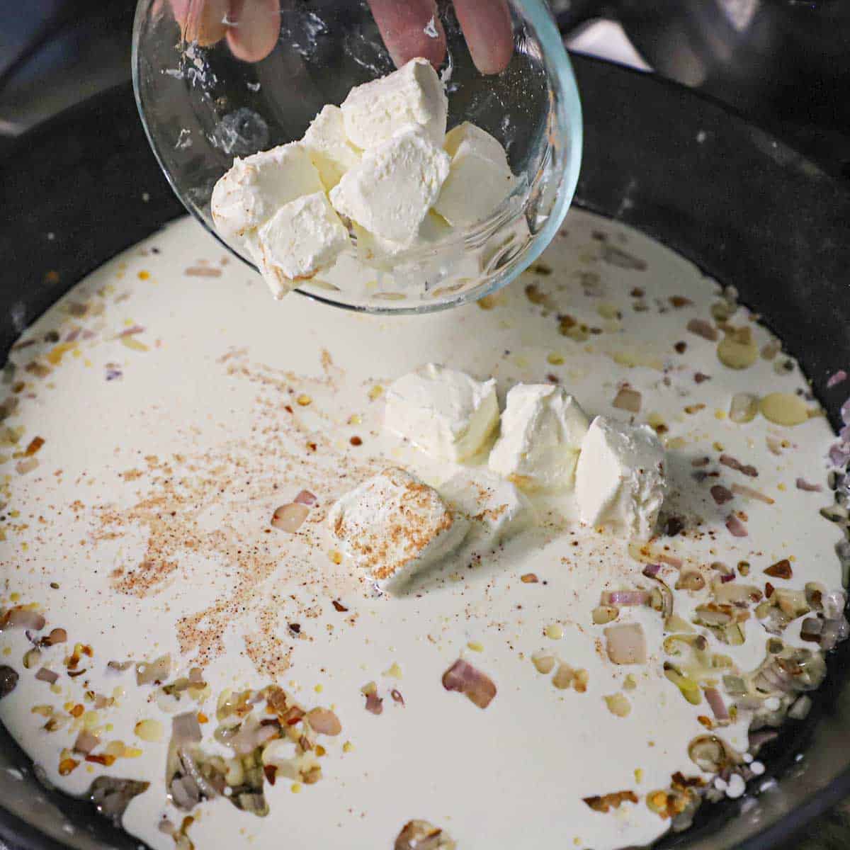 A person transferring cubed cream cheese from a small glass bowl into a cast-iron skillet filled with simmering cream with sautéed shallots and garlic.