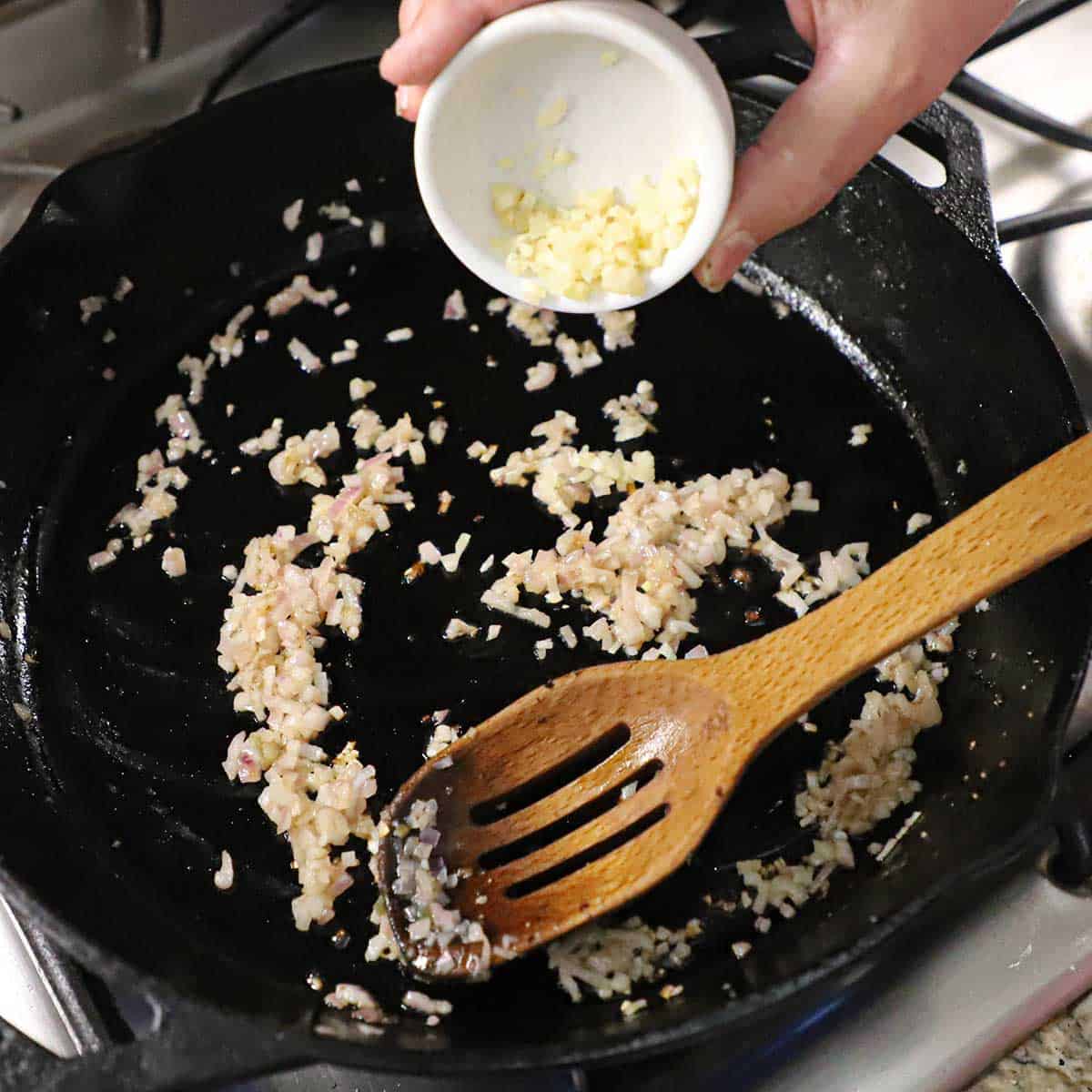 A person dumping minced garlic into a cast-iron skillet that contains finely chopped shallot that is being sautéed in butter in a cast-iron skillet.
