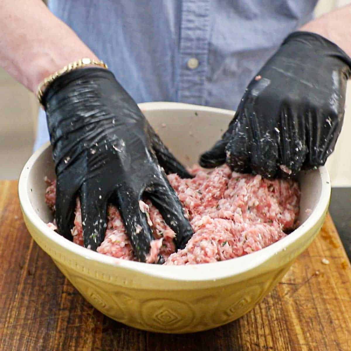 A person using his gloved hands to mix an Italian sausage meat mixture in a large ceramic bowl on a wooden cutting board.