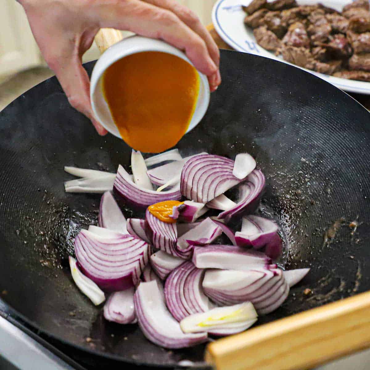 A person pouring aji amarillos paste from a small white bowl into a wok that is filled with wedges of red onion.